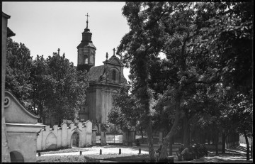 ML/H/593/210 Fotografia czarnobiała z widokiem dawnych zabudowań pomisjonarskich (obecnego Seminarium Duchownego) w Lublinie. Na pierwszym planie po lewej stronie widoczny fragment dworu Bentkowskich, skwer przed dworem (ob. Skwer im. bł. ks. Antoniego Zawistowskiego) oraz chodnik przy dawnej ul. Zamojskiej (ob. ul. Wyszyńskiego) z gęsto posadzonymi w rzędzie drzewami liściastymi, zajmującymi niemal całą prawą stronę fotografii. Pod drzewami na ławkach przy chodniku siedząca grupa dwunastu lub trzynastu zajętych rozmową mężczyzn w garniturach i czapkach na głowach. W tle neogotycki mur seminarium z bramą wjazdową w stylu eklektycznym. W murze łukowate wejście z otwartą metalową furtką. Nad wejściem napis „SEMINARIUM DUCHOWNE”. Przy murze siedząca starsza kobieta ubrana w ciemną suknię i chustę na głowie. Wzdłuż muru klasztornego wybrukowana ulica Misjonarska. Za murem na wzniesieniu Żmigród późno barokowy kościół pw. Przemienienia Pańskiego. Po lewej stronie na terenie seminarium drzewa liściaste zasłaniające część kościoła.