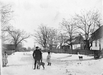 Fotografia czarno-biała przedstawia zimowy krajobraz wiejski. Na pierwszym planie starszy mężczyzna z psem oraz towarzysząca mu grupa dzieci. Po obydwu stronach ośnieżonej drogi znajdują się drewniane zabudowania oraz wysokie gołe drzewa. W oddali, na końcu drogi widoczna wieża kościoła.  