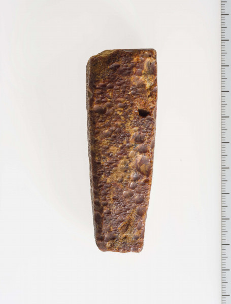 Surowiec bursztynowy, wstępnie obrobiony w formie wydłużonego trapezu. Surowiec w całości pokryty korą, widoczne ślady obróbki mechanicznej fragmentów płaszczyzn.