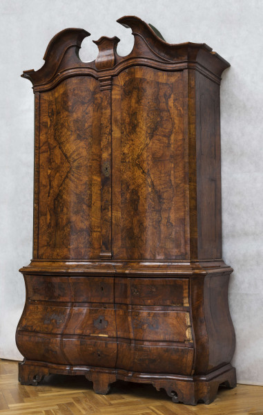 Szafa dwuczęściowa. Podstawa w formie trzyszufladowej komody, wsparta na czterech kostkowych nogach z wolutowymi ozdobami i niewielkim fartuchem. Szafa u góry obwiedziona profilowaną listwą, fronty szuflad wklęsło-wypukłe. Górna część węższa i cofnięta, dwudrzwiowa, zwieńczona wydatnym gzymsem wolutowym typu 