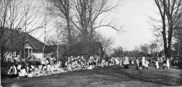 Fotografia czarno-biała przedstawia grupę mieszkańców wsi ubranych w stroje ludowe, którzy na klęczkach oczekują na obrzęd poświęcenia pokarmów. W oddali widoczne drewniane zabudowania wiejskie.