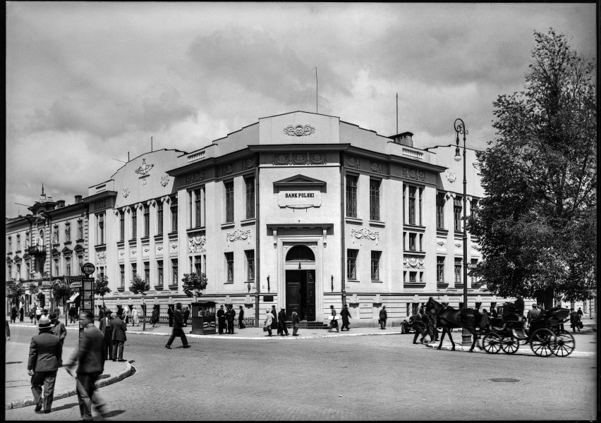 Fotografia czarnobiała z widokiem narożnego budynku Banku Polskiego na skrzyżowaniu ulic Krakowskie Przedmieście i 3 Maja w Lublinie. 
Zabudowa będąca częścią dziewiętnastowiecznego kompleksu Komisji Obwodu Lubelskiego. W centrum dwukondygnacyjny, otynkowany budynek dawnej Komory Celnej, kryty spadzistym dachem, z wejściem z poziomu ulicy. Na elewacji tablice informacyjne z nazwą instytucji mającej siedzibę w gmachu. Na szczytach widoczne fragmenty dekoracji. Z prawej strony kadru wysokie rozłożyste drzewo, pod którym stoi zaparkowana dorożka konna. Obok drzewa uliczna latarnia. Na chodnikach tłum przechodniów. Z lewej strony, na pierwszym planie, stoją dwaj mężczyźni w ciemnych garniturach. U dołu fotografii widoczna brukowana nawierzchnia. Ujęcie wykonane od strony rozwidlenia ulic Krakowskie Przedmieście oraz 3 Maja. 