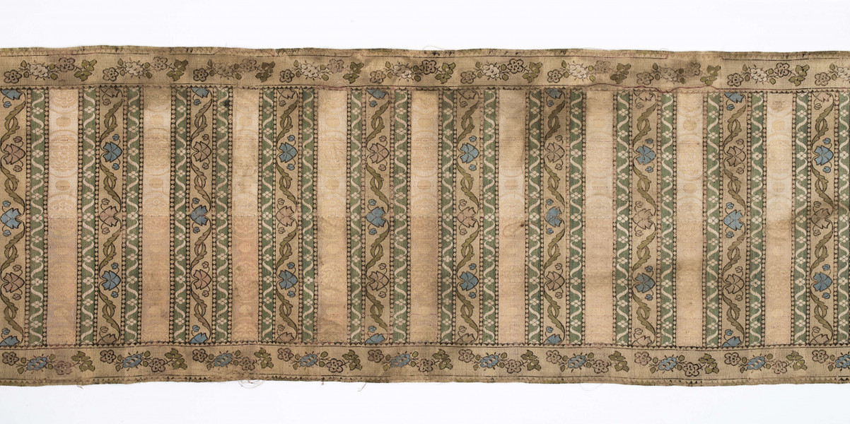 Pas 4-stronny. 
Ornament: głowy - krzaczki kwiatowe w wazonach; środek - poprzeczne pólka w układzie a,b,a,c (a - motyw wijacego sie wężyka, b - medaliony z rozetami, c - wić kwiatowa), obrzeża pionowe z gałązkami kwiatowymi, poziome - jak pólka c.
Koloryt: prawa strona - tło słomkowe, nieznacznie zróżnicowane w każdej połowie, zielen, błękit, żółty; lewa strona - tło brązowe głów i obrzeżeń, ornament - tło słomkowe, wzór j.w..
Fredzla doszyta jedwabna, pleciona, słomkowożółta