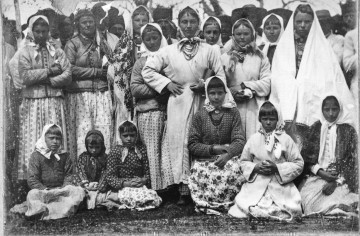 Fotografia czarno-biała przedstawia grupę w strojach ludowych. W pierwszym rzędzie dzieci siedzące na trawie, w drugim rzedzie stoją kobiety, za nimi mężczyźni. 