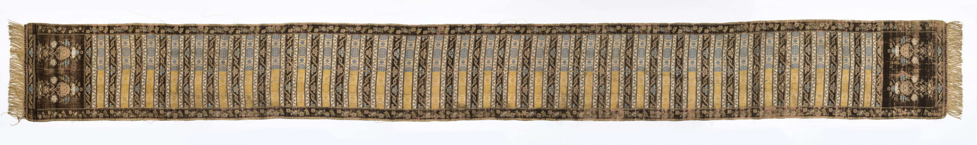 Pas 4-stronny. 
Ornament: głowy - krzaczki kwiatowe w wazonach; środek - poprzeczne pólka w układzie a,b,a,c (a - motyw wijacego sie wężyka, b - medaliony z rozetami, c - wić kwiatowa), obrzeża pionowe z gałązkami kwiatowymi, poziome - jak pólka c.
Koloryt: prawa strona - tło słomkowe, nieznacznie zróżnicowane w każdej połowie, zielen, błękit, żółty; lewa strona - tło brązowe głów i obrzeżeń, ornament - tło słomkowe, wzór j.w..
Fredzla doszyta jedwabna, pleciona, słomkowożółta