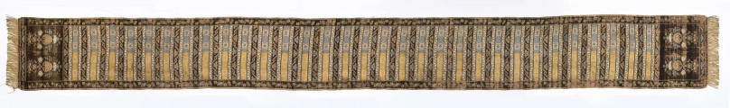 Pas 4-stronny. 
Ornament: głowy - krzaczki kwiatowe w wazonach; środek - poprzeczne pólka w układzie a,b,a,c (a - motyw wijacego sie wężyka, b - medaliony z rozetami, c - wić kwiatowa), obrzeża pionowe z gałązkami kwiatowymi, poziome - jak pólka c.
Koloryt: prawa strona - tło słomkowe, nieznacznie zróżnicowane w każdej połowie, zielen, błękit, żółty; lewa strona - tło brązowe głów i obrzeżeń, ornament - tło słomkowe, wzór j.w..
Fredzla doszyta jedwabna, pleciona, słomkowożółta