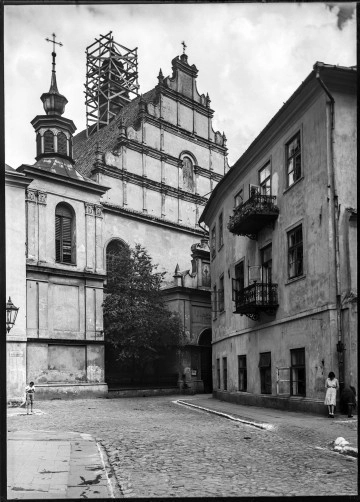 Fotografia czarnobiała z widokiem kościoła Dominikanów przy ul. Złotej w Lublinie. Ujęcie zamykające perspektywę ulicy Złotej. W centralnej części renesansowy szczyt kościoła Dominikanów z malowidłem przedstawiającym św. biskupa Stanisława, patrona świątyni. Za szczytem, wokół wieżyczki, wznosi się drewniane rusztowanie. Z prawej strony widoczna bryła dwupiętrowej kamienicy z balkonami oraz oknami na niskim parterze. Z lewej na fasadzie budynku oszklona latarnia uliczna oraz tablica urzędowa z nazwą ulicy. W głębi wejście do kościoła z wiszącą obok klepsydrą. Na lewo od wejścia ogrodzony żelazną balustradą niewielki parkan z rozłożystym drzewem. Nad portalem, na zwieńczeniu kruchty, malowidło z wizerunkiem relikwii Drzewa Krzyża Świętego. Po obu stronach brukowanej ulicy z bielonymi krawężnikami stoją postacie. 