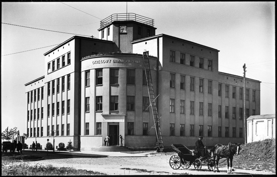 Fotografia czarnobiała z widokiem dawnego Urzędu Ziemskiego (ob. siedziby Wielonarodowej Brygady Litewsko-Polsko-Ukraińskiej i Muzeum Martyrologii „Pod Zegarem”). Na pierwszym planie, w prawym dolnym rogu fotografii, stoi dorożka konna z siedzącym woźnicą ze spuszczoną głową. Druga dorożka widoczna w lewym dolnym rogu w głębi ulicy Uniwersyteckiej. Za nimi, u zbiegu obecnych ulic Radziszewskiego i Uniwersyteckiej, budynek narożny w stylu modernistycznym. W parterowych oknach zamontowane metalowe ażurowe kraty. Widać doprowadzone przewody podłączone na dachu. Na szczycie wieży, na fasadzie budynku, kwadratowy zegar. O front, po prawej stronie od wejścia do gmachu, oparta długa drabina, zabezpieczona deskami wpartymi o okna parteru i pierwszego piętra. Na schodach kilku mężczyzn. Przed budynkiem wzdłuż ulicy Uniwersyteckiej, między chodnikiem a ścianą budynku, drzewa i krzewy. Ulice wyłożone brukiem, trawniki na pierwszym planie zaniedbane. W prawym dolnym rogu fragment otynkowanego i pobielonego muru na nasypie ziemnym i drewniany słup z przewodami elektrycznymi (ob. nieistniejące).