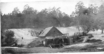 Fotografia czarno-biała przedstawia krajobraz wiejski z drewnianym młynem na pierwszym planie. W tle liczne drzewa. 