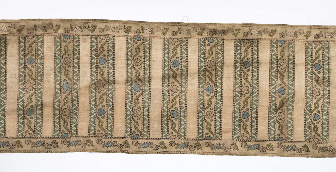 Pas 4-stronny. 
Ornament: głowy - krzaczki kwiatowe w wazonach; środek - poprzeczne pólka w układzie a,b,a,c (a - motyw wijacego sie wężyka, b - medaliony z rozetami, c - wić kwiatowa), obrzeża pionowe z gałązkami kwiatowymi, poziome - jak pólka c.
Koloryt: prawa strona - tło słomkowe, nieznacznie zróżnicowane w każdej połowie, zielen, błękit, żółty; lewa strona - tło brązowe głów i obrzeżeń, ornament - tło słomkowe, wzór j.w..
Fredzla doszyta jedwabna, pleciona, słomkowożółta