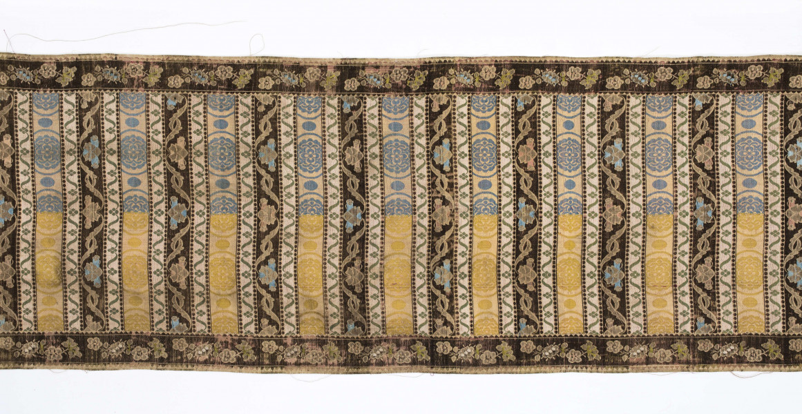 Pas 4-stronny. 
Ornament: głowy - krzaczki kwiatowe w wazonach; środek - poprzeczne pólka w układzie a,b,a,c (a - motyw wijacego sie wężyka, b - medaliony z rozetami, c - wić kwiatowa), obrzeża pionowe z gałązkami kwiatowymi, poziome - jak pólka c.
Koloryt: prawa strona - tło słomkowe, nieznacznie zróżnicowane w każdej połowie, zielen, błękit, żółty; lewa strona - tło brązowe głów i obrzeżeń, ornament - tło słomkowe, wzór j.w..
Fredzla doszyta jedwabna, pleciona, słomkowożółta