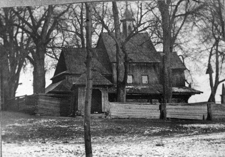 Fotografia czarno-biała przedstawia drewnianą cerkiew unicką otoczoną drewnianym ogrodzeniem. Ziemia częściowo pokryta jest śniegiem, a wokół budowli wznoszą się nagie drzewa. 