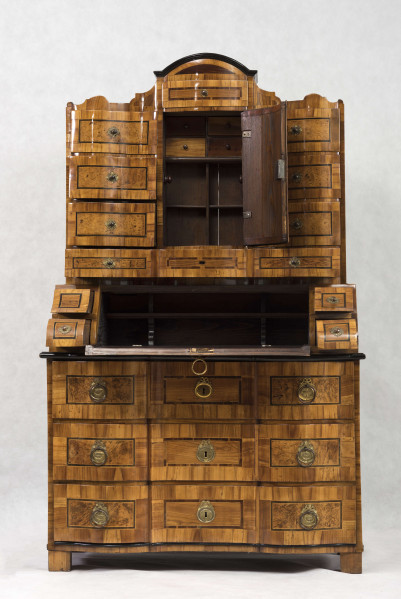 Sekretera trójkondygnacyjna. Podstawa w formie trzyszufladowej komody,wspartej na czterech kostkowych nogach. Środkowa część z prostokątnym skośnym, odchylnym blatem, obwiedziona profilowaną listwą, fronty szuflad wklęsło-wypukłe. Po bokach ukośnego blatu po dwie szufladki. Nastawa cofnięta z dwoma rzędami szufladek po bokach (po 4); część środkowa wyższa, w niej półkoliście wypukłe drzwiczki i dwie szufladki nad i pod drzwiczkami. Fronty szuflad faliste jak i wykrój profilowanego zwieńczenia. Powierzchnia intarsjowana, dekorowana fornirem: na drzwiczkach kompozycja w formie gałązki z kwiatami, na blacie symetrycznie ułożone dwa ornamenty liściasto-wolutowe,  na szufladach poziome prostokątny ujęte w ciemne ramki. Nakładane brązy:  szyldziki do kluczy, antabki i kuliste uchwyty. 
