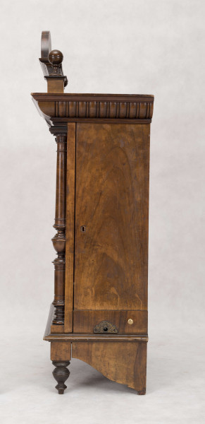 Pozytywka w szafce eklektycznej, drewnianej pokrytej okleiną z drewna orzechowego. Szafka prostokątna, pionowa, z ozdobnym zwieńczeniem, na niskich nóżkach, na froncie drzwiczki. Pośrodku zwieńczenia prostokątny cokolik z muszlą, po bokach kuliste sterczynki na sześciennych podstawach. Gzyms wieńczący górą wysunięty z rytowanym pośrodku  napisem 