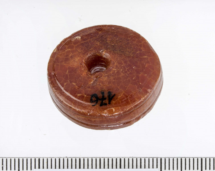 Paciorek typu II, tarczowaty, wypukły. Obie płaszczyzny paciorka zdobione pojedyńczymi liniami rytymi. Bok paciorka zdobiony parą dookolnych bruzd. Widoczne ślady toczenia.