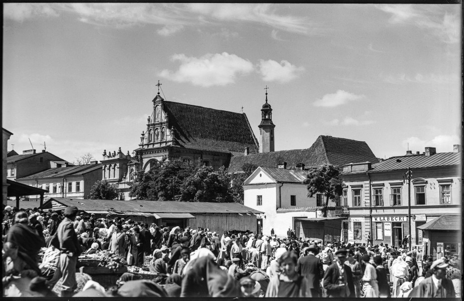 Fotografia czarnobiała z widokiem targu przy ul. Świętoduskiej w Lublinie z kościołem Karmelitów w tle.
Teren targowiska miejskiego na Górce Świętoduskiej – między ulicami Świętoduską a Nową (obecnie Lubartowską). Na pierwszym planie stragany oraz piętrowe zabudowania jatek. Wokół tłum ludzi – sprzedawców i kupujących. W głębi widoczna bryła kościoła Karmelitów, zza której wyłania się wieżyczka zabudowań klasztornych. Po obu stronach świątyni rzędy kamienic z reklamami sklepów i zakładów usługowych. 