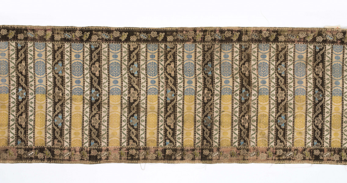 Pas 4-stronny. 
Ornament: głowy - krzaczki kwiatowe w wazonach; środek - poprzeczne pólka w układzie a,b,a,c (a - motyw wijacego sie wężyka, b - medaliony z rozetami, c - wić kwiatowa), obrzeża pionowe z gałązkami kwiatowymi, poziome - jak pólka c.
Koloryt: prawa strona - tło słomkowe, nieznacznie zróżnicowane w każdej połowie, zielen, błękit, żółty; lewa strona - tło brązowe głów i obrzeżeń, ornament - tło słomkowe, wzór j.w..
Fredzla doszyta jedwabna, pleciona, słomkowożółta