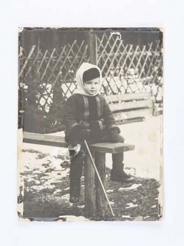 Adam Żeromski w góralskim stroju (kapelusz,chusta, kożuszek obszyty baranim futrem) oparty na laseczce siedzi na wyrwanych korzeniach drzewa, na tle lasu.