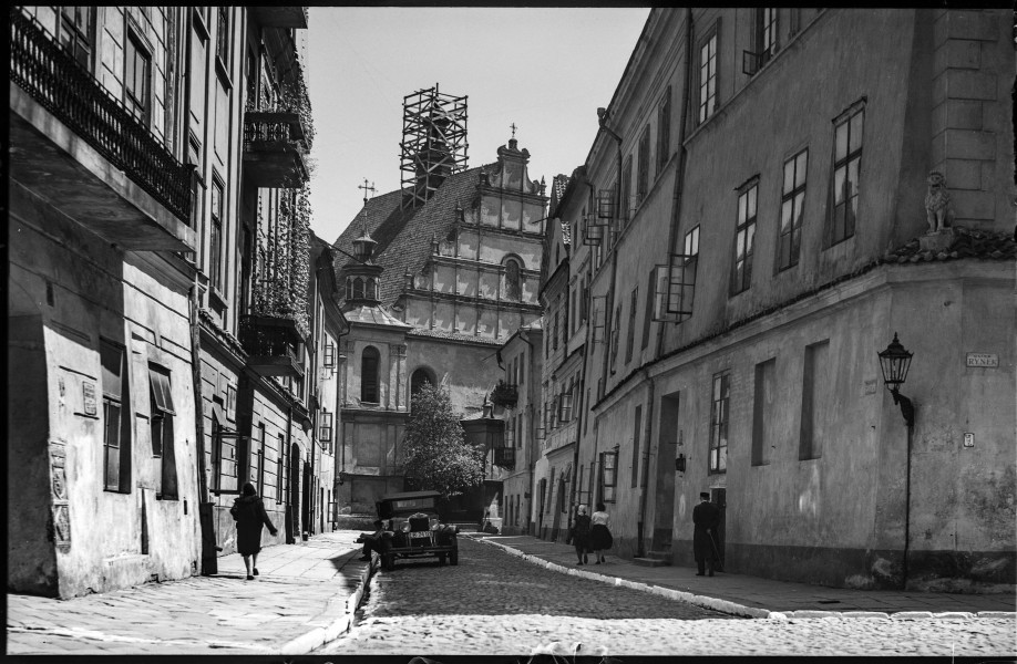 Fotografia czarnobiała z widokiem ulicy Złotej w Lublinie z kościołem Dominikanów w głębi.
Ujęcie zamykające perspektywę ulicy Złotej od strony skrzyżowania z ulicą Rynek. W głębi centralnej części kadru widoczny renesansowy szczyt kościoła Dominikanów z malowidłem we wnęce przedstawiającym św. biskupa Stanisława, patrona świątyni. Za szczytem, wokół wieżyczki, wznosi się drewniane rusztowanie. Po lewej od wejścia rozłożyste drzewo. Z prawej strony widoczne fasady kamienic. Narożna kamienica „Pode Lwami” (Rynek 9) z figurką kamiennego lwa ustawioną na gzymsie oddzielającym parter od pierwszej kondygnacji. Poniżej lwa przeszklona uliczna latarnia oraz tablica urzędowa z nazwą ulicy. Z lewej strony fasady kamienic z balkonami z pnączami roślin. Na rogu fragment Kamienicy Lubomelskich (Rynek 8) z widocznym wmurowanym kamiennym gmerkiem z herbem Zadora. Otwarte skrzydła okien uchylone na zewnątrz. Po obu stronach brukowanej ulicy z bielonymi krawężnikami idący przechodnie (w tym Żyd w chałacie) odwróceni tyłem do obiektywu. Na pierwszym planie zaparkowany samochód osobowy z kierowcą (?) siedzącym na zewnątrz z nogami opartymi o krawężnik. 
