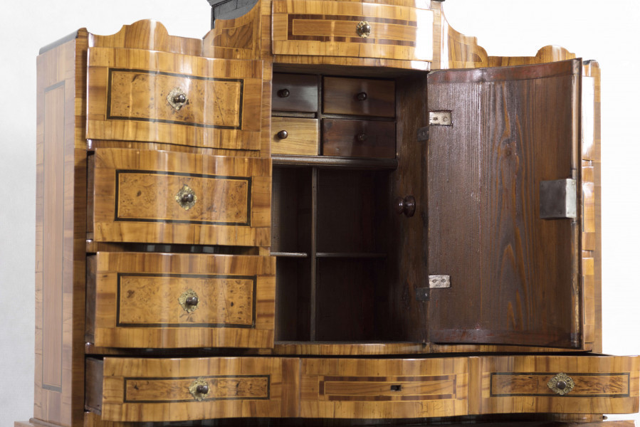 Sekretera trójkondygnacyjna. Podstawa w formie trzyszufladowej komody,wspartej na czterech kostkowych nogach. Środkowa część z prostokątnym skośnym, odchylnym blatem, obwiedziona profilowaną listwą, fronty szuflad wklęsło-wypukłe. Po bokach ukośnego blatu po dwie szufladki. Nastawa cofnięta z dwoma rzędami szufladek po bokach (po 4); część środkowa wyższa, w niej półkoliście wypukłe drzwiczki i dwie szufladki nad i pod drzwiczkami. Fronty szuflad faliste jak i wykrój profilowanego zwieńczenia. Powierzchnia intarsjowana, dekorowana fornirem: na drzwiczkach kompozycja w formie gałązki z kwiatami, na blacie symetrycznie ułożone dwa ornamenty liściasto-wolutowe,  na szufladach poziome prostokątny ujęte w ciemne ramki. Nakładane brązy:  szyldziki do kluczy, antabki i kuliste uchwyty. 
