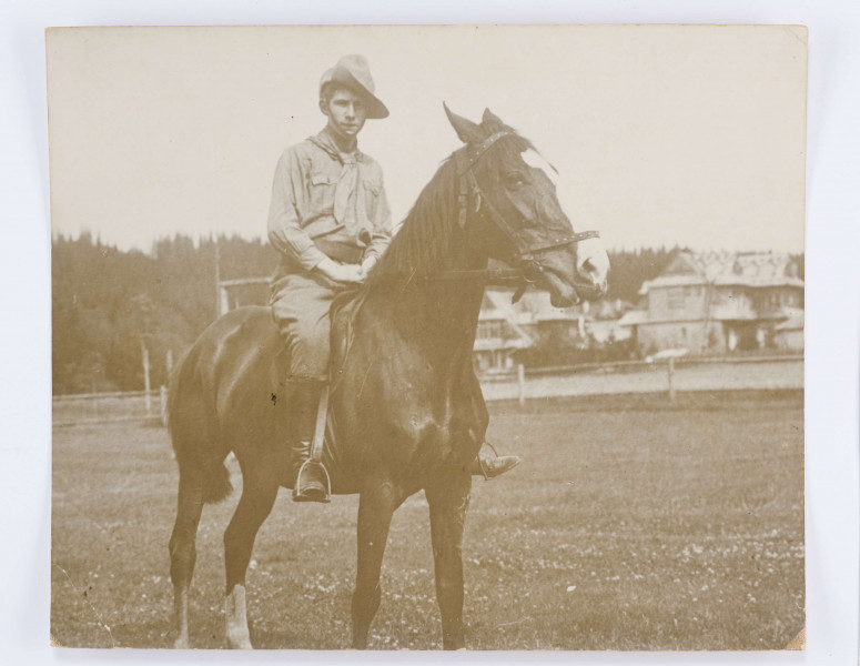 Fotografia Adama Żeromskiego na koniu. Adam w stroju skautowskim (bryczesy, bluza z zawiązaną chusta pod brodą) w kapeluszu na głowie siedzi na łysce w siodle. Nogi w wysokich butach trzyma w strzymionach. Fotografia wykonana na tle lasu. Po prawej stronie widac budynki (pensjonat).