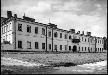 Fotografia czarnobiała przedstawiająca fasadę główną Katolickiego Uniwersytetu Lubelskiego (dawne koszary rosyjskie) od strony alei Racławickich. Widoczny pobielony gmach jednopiętrowy z dwoma rzędami okien i fragmentem skrzydła wschodniego, do którego przylega murek sięgający do połowy pierwszego rzędu okien, w środkowej części, odsuniętej w tył w stosunku do flankujących ją ryzalitów, widoczne wejście pod półokrągłym sklepieniem. Nad wejściem pojedynczy metalowy balkon. Przed gmachem zaniedbany trawnik i fragment chodnika z płyt betonowych i częściowo z bruku. W lewym dolnym rogu i w oddali po prawej stronie widoczne dojścia do gmachu od strony alei.