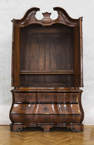 Szafa dwuczęściowa. Podstawa w formie trzyszufladowej komody, wsparta na czterech kostkowych nogach z wolutowymi ozdobami i niewielkim fartuchem. Szafa u góry obwiedziona profilowaną listwą, fronty szuflad wklęsło-wypukłe. Górna część węższa i cofnięta, dwudrzwiowa, zwieńczona wydatnym gzymsem wolutowym typu 