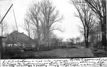 Fotografia czarno-biała przedstawia krajobraz wiejski z drewnianą zabudową i ogrodzeniem, żurawiem studziennym i drzewami.