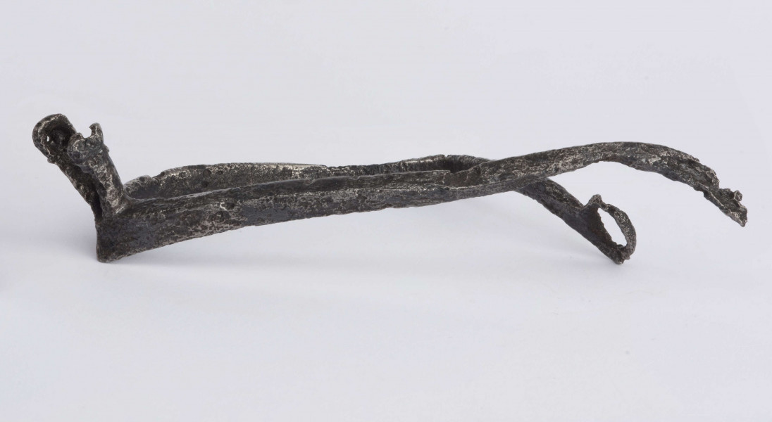 Ostroga żelazna. Kabłąk o trójkątnym przekroju poprzecznym, zakończony zaczepem w kształcie kółka. Bodziec (przedłużenie kabłąka) zakończone widełkami. Jedno z ramień widełek na wysokości otworu złamane, drugie zachowane w całości z otworem dla umocnowania gwiazdki. Opisywany egzemplarz nie posiadagwiazdki, kabłąk z zaczepem jest zgięty, drugi bez zaczepu.
