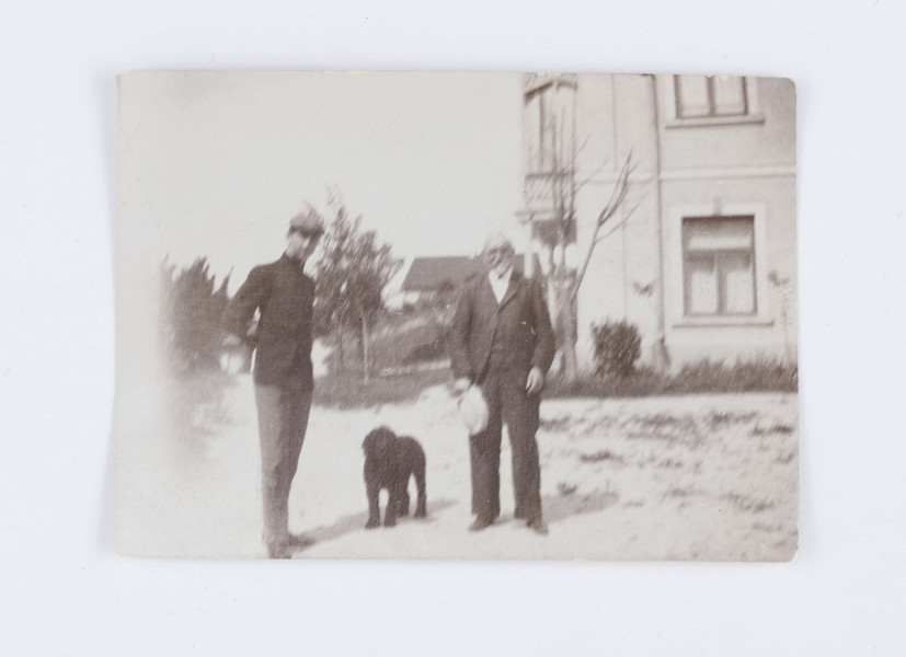 Fotografia przedstawia Emila Trembińskiego z nieznanym mężczyzną. Emil stoi po lewej stronie, ubrany jest w czarny surdut oraz szare spodnie, a na głowie znajduje się szary kaszkiet. Ręce trzyma za plecami. Po prawej stronie stoi starszy mężczyzna ubrany w ciemną marynarkę, spodnie, kamizelkę oraz białą koszulę. W prawej ręce trzyma biały kapelusz. Między mężczyznami widać czarnego psa. W tle, po prawej stronie, widać fragment  murowanego, jasnego, piętrowego budynku. Po lewej stronie znajduje się kilka drzew i  krzaków. W oddali widać zarysy pojedynczej chaty. Na odwrocie znajduje się odręczny napis: „Nałęczów/ Emil Trembiński/ R. 1900”.