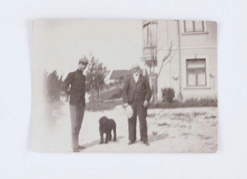 Fotografia przedstawia Emila Trembińskiego z nieznanym mężczyzną. Emil stoi po lewej stronie, ubrany jest w czarny surdut oraz szare spodnie, a na głowie znajduje się szary kaszkiet. Ręce trzyma za plecami. Po prawej stronie stoi starszy mężczyzna ubrany w ciemną marynarkę, spodnie, kamizelkę oraz białą koszulę. W prawej ręce trzyma biały kapelusz. Między mężczyznami widać czarnego psa. W tle, po prawej stronie, widać fragment  murowanego, jasnego, piętrowego budynku. Po lewej stronie znajduje się kilka drzew i  krzaków. W oddali widać zarysy pojedynczej chaty. Na odwrocie znajduje się odręczny napis: „Nałęczów/ Emil Trembiński/ R. 1900”.