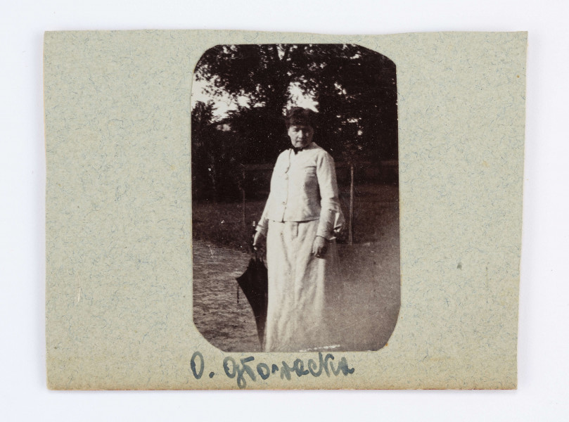 Fotografia naklejona na kawałek tektury. Oktawia Głowacka stoi na ścieżce, ubrana w białą spódnicę i żakiet. W prawej ręce trzyma czarną parasolkę. Za plecami Oktawii można zauważyć kilka drzew. Pod fotografią znajduje się podpis: O. Głowacka.
