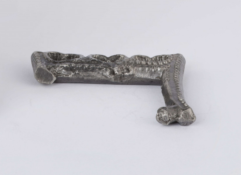 Sprzączka do pasa zachowana fragmentarycznie wykonana ze srebra, zdobiona . Ramka prostokątna. Przednia część ramki zdobiona trzema wypukłymi sercowatymi ornamentami z wyciętymi środkami w kształcie półksiężyca. Wszystkie wypukłości wyżej opisanego ornamentu oraz krawędzie sprzączki zdobione drobnym granulatem. Surowiec - srebro. Wymiary: średnia grubość 0,4; wymiary boków ramki 3,4 i 3,2 cm. Stan zachowania dobry. 