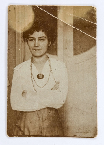 MC/M/845/ML Fotografia w formie stojącego prostokąta, przedstawiająca Katarzynę Czechowiczówną (3/4 ujętej postaci) na tle białych, oszklonych drzwi. Katarzyna stoi nieco po lewej stronie, z załozonymi rękami. Ubrana jest w białą bluzkę i spódnicę. Na szyi ma drobne korale, do bluzki przypięta ciemna broszka. Włosy ciemne, kręcone, upięte z tyłu, ciemne, grube brwi, na twarzy lekki uśmiech. Fotografia monochromatyczna, sepia. Powierzchnia papieru gładka, lekko świecąca. Nad głową postaci dwie białe plamy. Na odwrocie, w górnej części ślady po kleju, nizej numer inwentarza napisany ołówkiem.