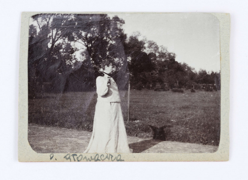 Fotografia przedstawia Oktawie Głowacką z psem. Kobieta siedzi na schodach, ubrana jest w długą spódnicę, czarny płaszcz i białą chustę na głowie. Na kolanach trzyma małego, brązowego psa. Z tyłu widać kobietę w kraciastej spódnicy, która trzyma w lewej ręce czarną laskę. Na prawo od Oktawii widać oparty o balustradę schodów czarny parasol. Zdjęcie przyklejone jest do tektury, u dołu której widniej odręczny podpis: O. Głowacka.