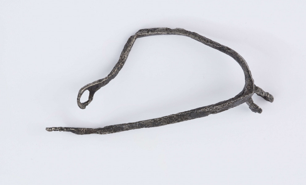 Ostroga żelazna. Kabłąk o trójkątnym przekroju poprzecznym, zakończony zaczepem w kształcie kółka. Bodziec (przedłużenie kabłąka) zakończone widełkami. Jedno z ramień widełek na wysokości otworu złamane, drugie zachowane w całości z otworem dla umocnowania gwiazdki. Opisywany egzemplarz nie posiadagwiazdki, kabłąk z zaczepem jest zgięty, drugi bez zaczepu.