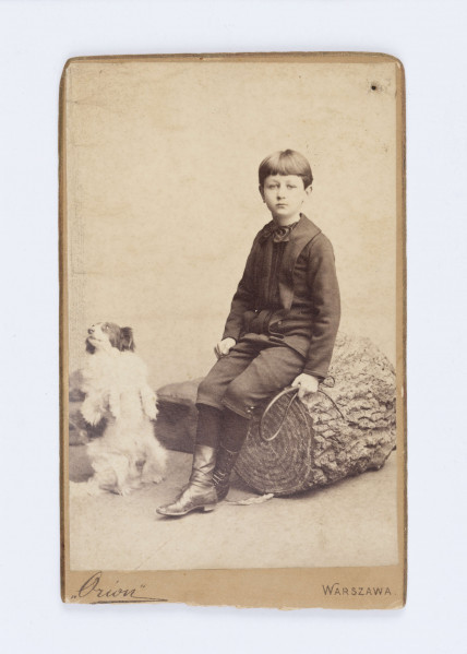 Fotografia przedstawia młodego Emila Trembińskiego. Chłopiec ubrany jest w ciemną marynarkę i takiego samego koloru spodnie, czarną koszulą, czarne pończochy oraz skórzane buty sięgające połowy łydki. W lewej dłoni trzyma mały bicz, prawą opiera o udo. Chłopiec siedzi na pniaku. Po jego prawej stronie widać biało-czarnego psa który stoi na dwóch tylnych łapkach.  W lewym, dolnym rogu znajduje się napis „Orion”. Na odwrocie, wzdłuż prawej krawędzi, widnie podpis wykonany ołówkiem: Emil Trembiński. Po środku znajduje się drukowana nazwa firmy „Orion”.