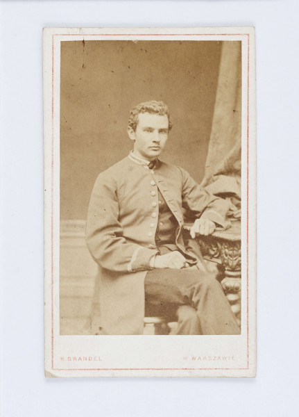 Fotografia przedstawia młodego Aleksandra Głowackiego. Pisarz siedzi na krześle, ubrany jest w długi, jasny surdut, na którym widać osiem jasnych guzików. Surdut jest zapięty  jedynie na pojedynczy guzik pod szyją. Pod spodem widać ciemną kamizelkę oraz tego samego koloru spodnie. Aleksander opiera się lewą ręką o bogato zdobiony stół. Całe zdjęcie jest otoczone białą ramką. Wzdłuż dolnej krawędzi znajduje się drukowany napis: K. Brandel w Warszawie. Na odwrocie, po środku znajduje się logo firmy która wykonała zdjęcie Logo składa się z odręcznego napisu K. Brandel z drukowanym, ciemnym podkreśleniem.