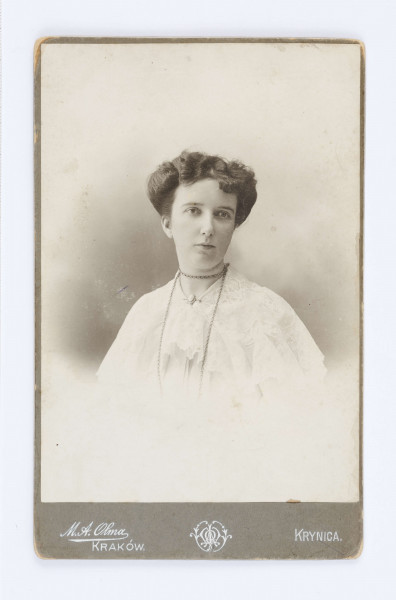 Fotografia przedstawia Julie Dąbrowską z Głoskowskich. Kobieta ubrana jest w biały, haftowany strój zapięty pod brodą przy pomocy broszki z jasnym kamieniem. Na szyi znajduje się długi łańcuszek, owinięty wokół szyi. Zdjęcie naklejone jest na firmową tekturę. W lewym, dolnym rogu znajduje się nazwa firmy która wykonała fotografię: M.A.Olma Kraków. W lewym dolnym rogu znajduje się napis: Krynica. Na odwrocie, wzdłuż lewej strony, znajduje się ilustracja przedstawiający kobietę, w długiej sukni z motywem kwiatowym, która w dłoniach trzyma lupę oraz fotografię z budynkiem, stojącą na tle kwiatu.  Po prawej stronie znajduje się drukowany napis z nazwą zakładu fotograficznego: „Zakład Artystyczno-fotograficzny M.A. Olmy w KRAKOWIE Podwale 14 i w Krynicy”.

