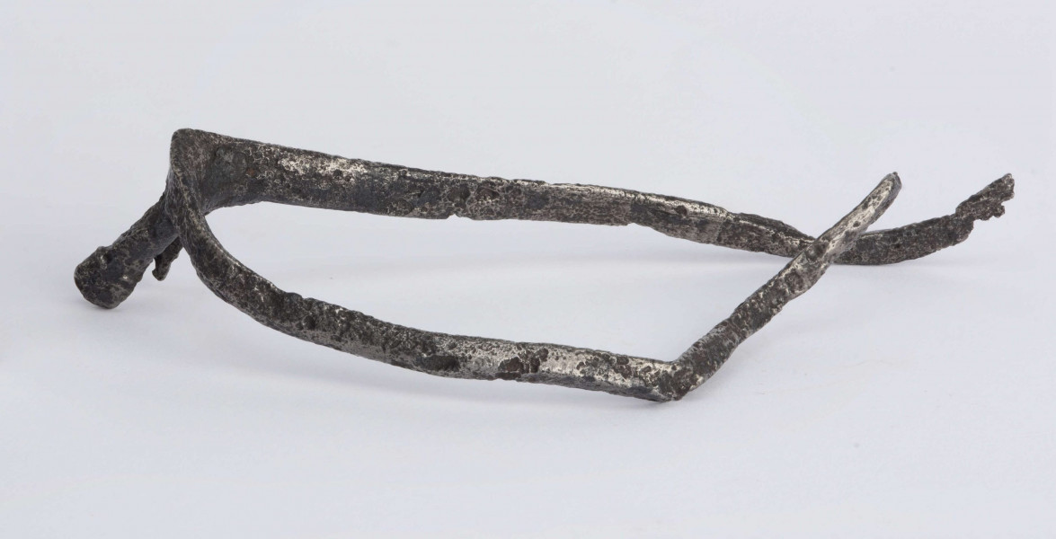 Ostroga żelazna. Kabłąk o trójkątnym przekroju poprzecznym, zakończony zaczepem w kształcie kółka. Bodziec (przedłużenie kabłąka) zakończone widełkami. Jedno z ramień widełek na wysokości otworu złamane, drugie zachowane w całości z otworem dla umocnowania gwiazdki. Opisywany egzemplarz nie posiadagwiazdki, kabłąk z zaczepem jest zgięty, drugi bez zaczepu.