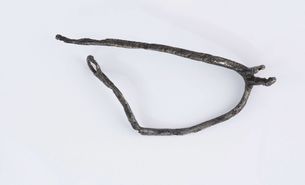 Ostroga żelazna. Kabłąk o trójkątnym przekroju poprzecznym, zakończony zaczepem w kształcie kółka. Bodziec (przedłużenie kabłąka) zakończone widełkami. Jedno z ramień widełek na wysokości otworu złamane, drugie zachowane w całości z otworem dla umocnowania gwiazdki. Opisywany egzemplarz nie posiadagwiazdki, kabłąk z zaczepem jest zgięty, drugi bez zaczepu.