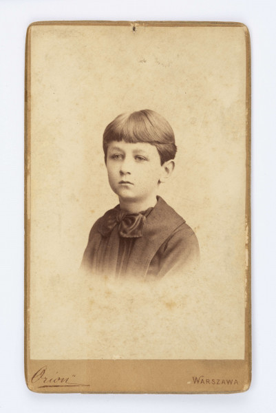 Fotografia portretowa Emila Trembińskiego. Chłopiec gładko uczesany, twarz skierowana w lewą stronę. Ubrany w ciemną koszulę z muszką pod brodą.  Pod fotografią znajduje się wydrukowany podpis: Orion Warszawa. Na odwrocie znajduje się przyklejony fragment białej kartki który zasłania wydrukowany napis. Z prawej strony widnieje napis wykonany ołówkiem: Emil Trembiński.