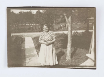 Fotografia przedstawia Marcjannę Karczewską. Kobieta ubrana jest w jasną, kraciastą, długą spódnicę oraz jasną koszulę w ciemne romby. Po lewej stronie znajduje się jasny chodnik. W tle widać gęsto rosnące drzewa oraz polanę porośniętą krzakami z białymi kwiatami. Po prawej stronie widać fragment, jasnego budynku. Na odwrocie widnieje odręczny napis wykonany długopisem: „ p. Marcysia, nasz gosposia w Nałęczowie”. 