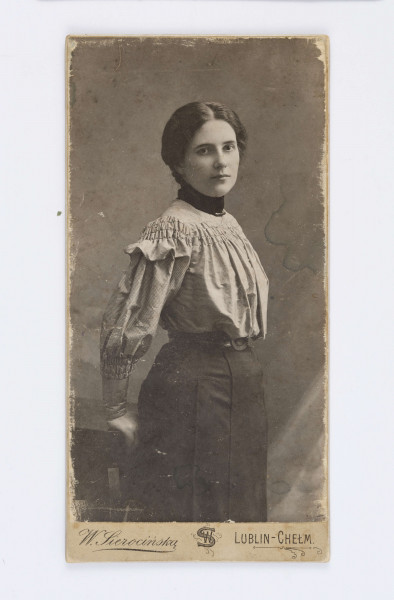 Fotografia przedstawia Janinę Juszkiewiczową z Głoskowskich. Ubrana jest w szarą koszulę, długą, ciemną spódnicę z czarnym paskiem, a pod szyją widać czarny kołnierzyk. Kobieta opiera się o drewniany stolik. W lewym, dolnym rogu znajduje się drukowany podpis autora zdjęcia: W. Sierocińska. W prawym dolnym rogu znajduje się napis: Chełm-Lublin. Na odwrocie znajduje się ilustracja z motywem kwiatowym. Obok znajduje się drukowany tekst: „W. Sierocińska/ Lublin-Chełm/ Niepogoda nie robi różnicy/ w zdjęciach/ Klisze przechowuje się/ na żądanie/ może mieć/ odbicia”.