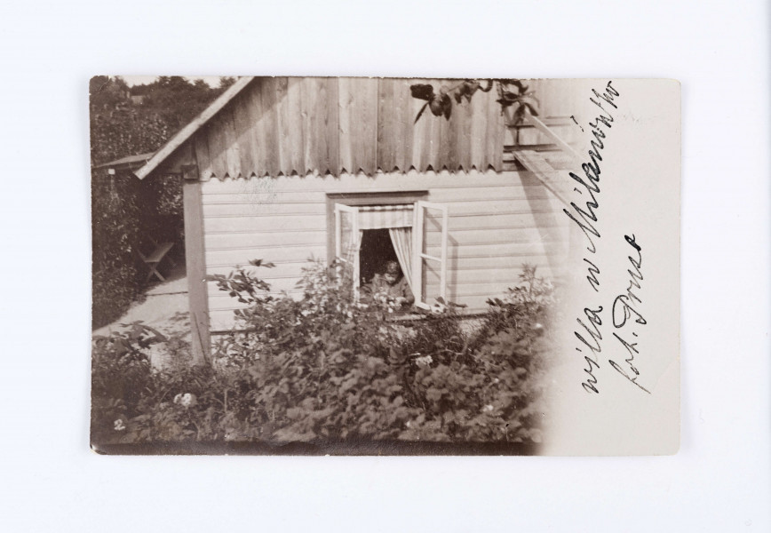 Fotografia przedstawia drewniany budynek, o spadzistym dachu. Na pierwszym planie widać otwarte, białe okno przez które wychyla się starsza kobieta. Widać również dwukolorowe firanki. Przed budynkiem widać krzewy i białe kwiaty. Po lewej stronie budynku, w tle, znajduje się drewniana ławeczka z oparciem. Prawa strona zdjęcia jest biała i znajduje się odręczny napis: „willa w Milanówku/ fot. Prus”.