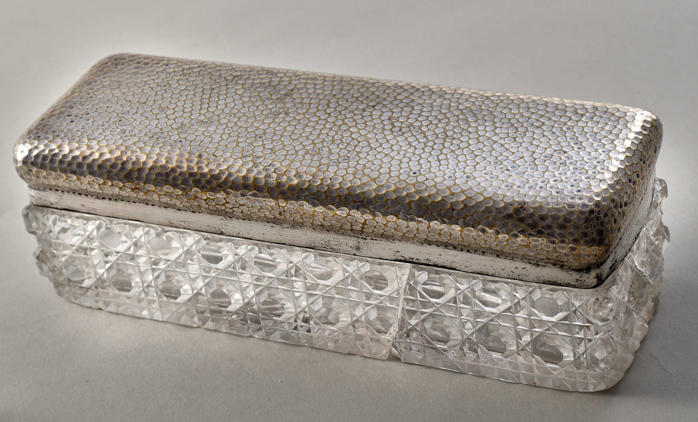 Na pierwszym planie widoczne jest pudełko z posrebrzanym spodem z wzorami oraz pokrywą.