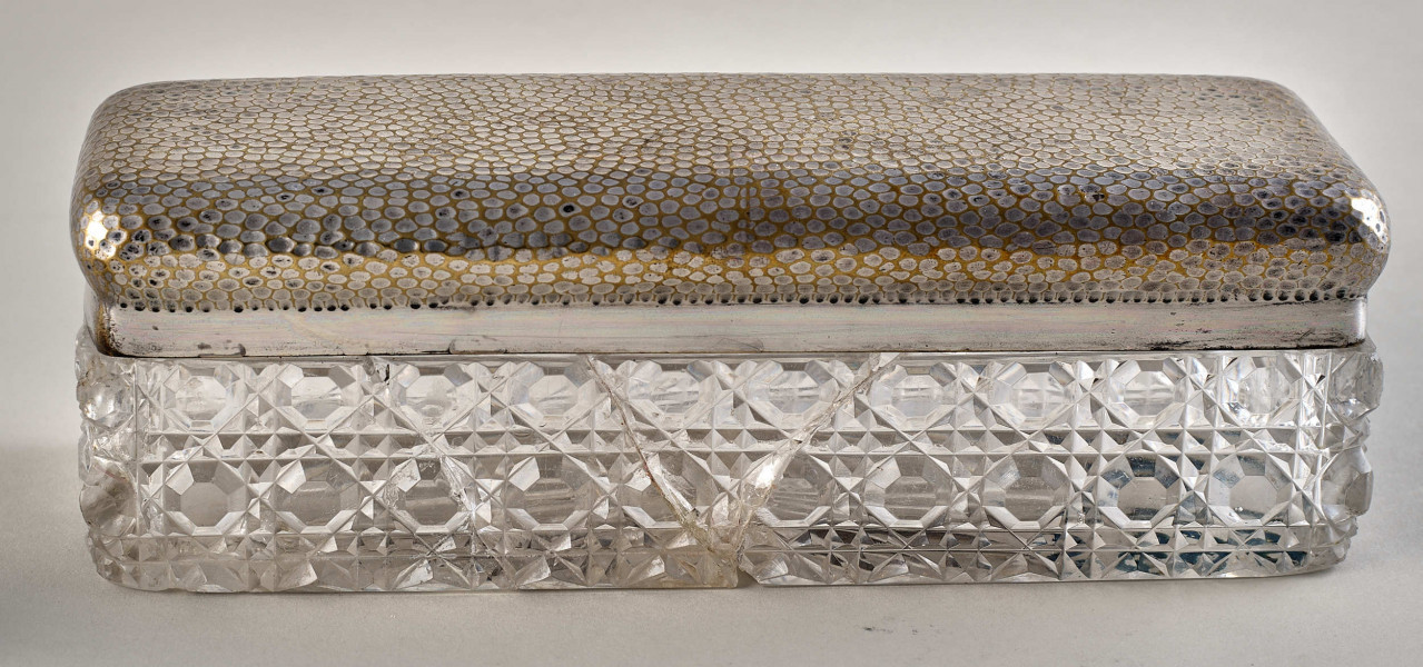 Na pierwszym planie widoczne jest pudełko z posrebrzanym spodem z wzorami oraz pokrywą.