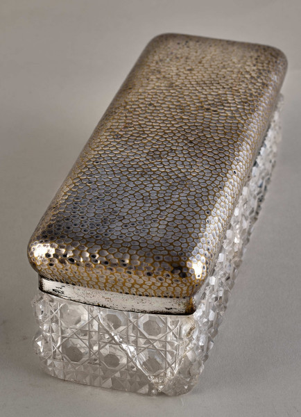 Na pierwszym planie widoczne jest pudełko z posrebrzanym spodem z wzorami oraz pokrywą.