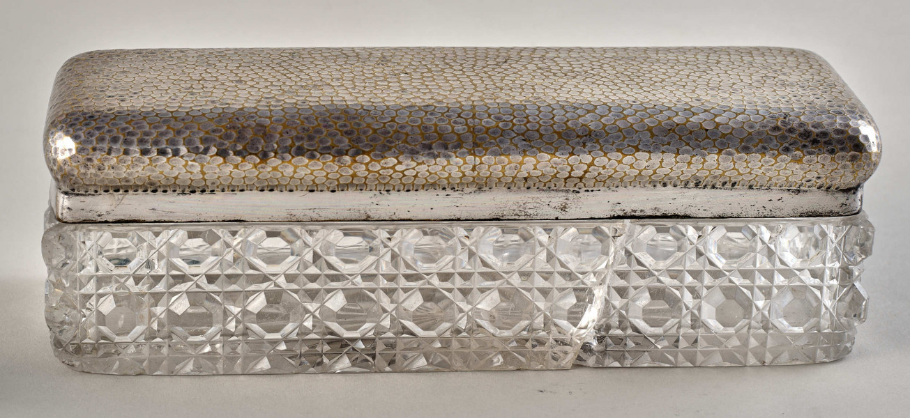 Na pierwszym planie widoczne jest pudełko z posrebrzanym spodem z wzorami oraz pokrywą.