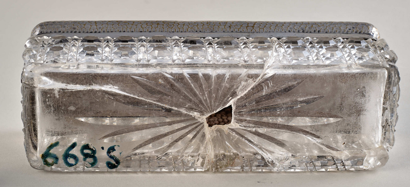 Na pierwszym planie widoczne jest pudełko z posrebrzanym spodem z wzorami oraz pokrywą. Spód